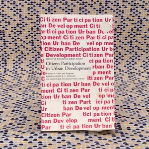Op de afbeelding: Een boek getiteld "Citizen Participation in Urban Development" met de ondertitel "Selected Readings Series Eight, Volume II - Cases and Programs" bewerkt door Hans A. C. Spiegel. De omslag van het boek is wit met rode tekst.