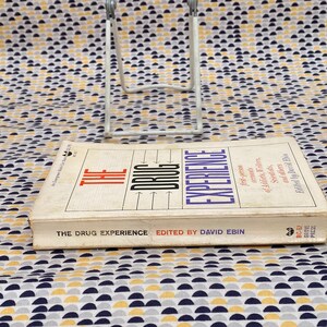 May include: A white book with the title "The Drug Experience" and the subtitle "First-person accounts of LSD, mescaline, marijuana, and others" by David Ebin. The book is on a white and yellow patterned surface.