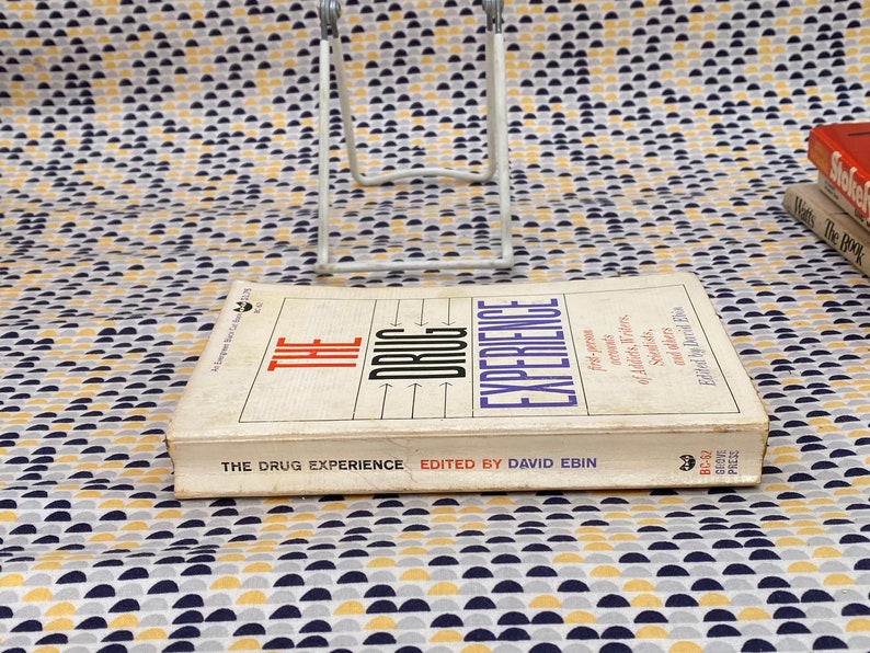 May include: A white book with the title "The Drug Experience" and the subtitle "First-person accounts of LSD, mescaline, marijuana, and others" by David Ebin. The book is on a white and yellow patterned surface.
