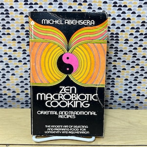 May include: A vintage cookbook titled "Zen Macrobiotic Cooking" with a black cover. The cover features a vibrant design with orange, yellow, and pink accents, and a yin-yang symbol. The book's title and author's name are prominently displayed.