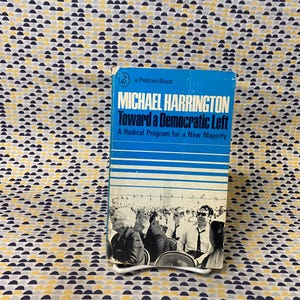 Op de afbeelding: Een blauw en wit paperback boek getiteld "Toward a Democratic Left: A Radical Program for a New Majority" door Michael Harrington. De omslag van het boek toont een zwart-witfoto van een groep mensen.