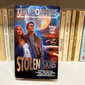 May include: A paperback book titled "Stolen Skies" by Tim Powers. The cover features a man and woman with a futuristic background. The book is displayed in front of a shelf of other books.