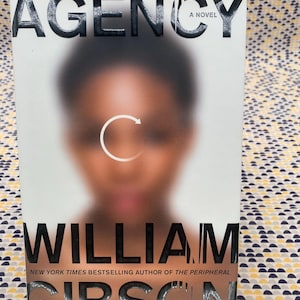 Puede incluir: Portada del libro "Agency" de William Gibson. El título y el nombre del autor están en letras negras grandes y en negrita. La portada presenta una imagen borrosa de una persona con una flecha circular blanca. El texto "A Novel" está encima de la imagen borrosa.