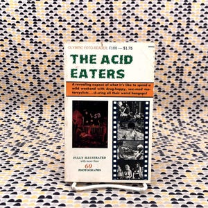 May include: A vintage paperback book titled "The Acid Eaters" with a white cover and green text. The cover features images and text describing a weekend with drug-happy motorcyclists. The book is fully illustrated with 60 photographs.