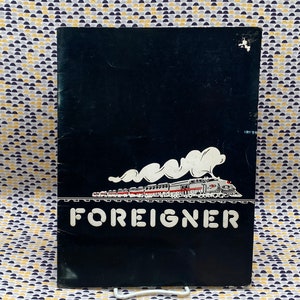 May include: Black and white album cover with a train and smoke in the background. The text "FOREIGNER" is printed in white letters on the front.