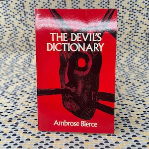 May include: A hardcover book titled "THE DEVIL'S DICTIONARY" by Ambrose Bierce. The cover is red with white text and a black illustration of a face. The book is standing upright against a patterned background.
