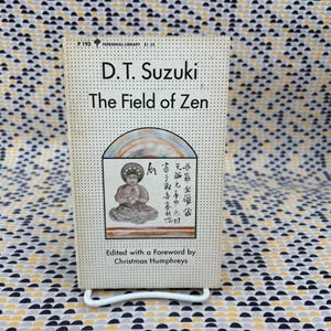 Pode incluir: Um livro de bolso vintage intitulado "The Field of Zen" de D.T. Suzuki. A capa apresenta uma ilustração de uma figura sentada e texto em inglês e outra língua. O livro é apoiado em um suporte de metal branco.