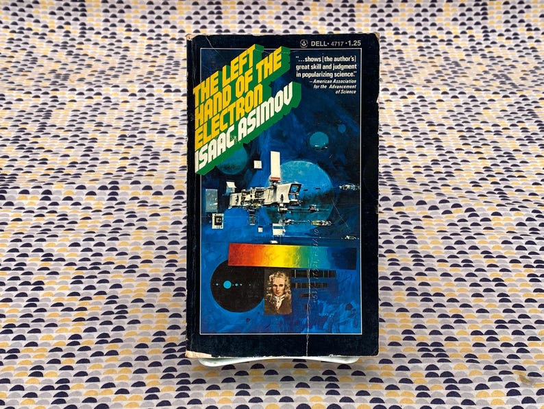 May include: A paperback book cover with a blue background and a space scene. The title is "The Left Hand of the Electron" by Isaac Asimov. The text on the cover reads "...shows the author's great skill and judgment in popularizing science." - American Association for the Advancement of Science.