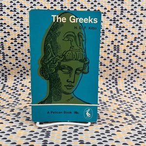 May include: A vintage paperback book titled "The Greeks" by H.D.F. Kitto. The book cover is teal with a green illustration of a classical bust. The book is a Pelican Book.