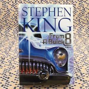 May include: A hardcover book titled "From A Buick 8" by Stephen King. The cover features a blue car with a bright headlight and the number 8. The book is set against a patterned background.