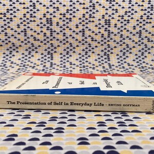 Puede incluir: Un libro de bolsillo titulado "The Presentation of Self in Everyday Life" de Erving Goffman. El libro tiene una cubierta blanca con detalles en rojo y azul. El libro est&aacute; sobre una superficie estampada.