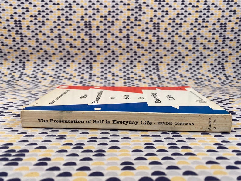 Puede incluir: Un libro de bolsillo titulado "The Presentation of Self in Everyday Life" de Erving Goffman. El libro tiene una cubierta blanca con detalles en rojo y azul. El libro est&aacute; sobre una superficie estampada.