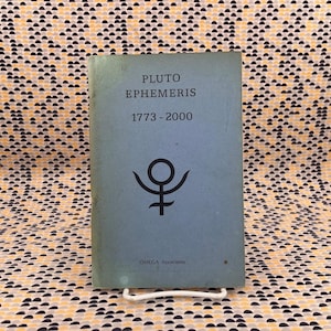 May include: A vintage book titled "PLUTO EPHEMERIS 1773-2000" with a black symbol on a light blue cover. The book is propped up on a white metal stand against a patterned background.