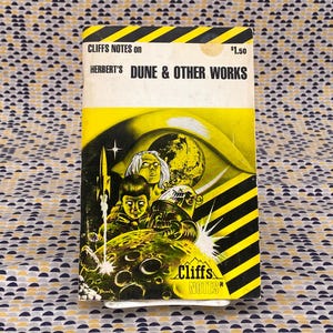 Może przedstawiać: Okładka książki Cliffs Notes w żółto-czarne paski z tytułem "Herbert's Dune & Other Works" i ceną 1,50 $. Okładka przedstawia ilustrację gigantycznego oka z planetą w środku, otoczonego trzema postaciami.