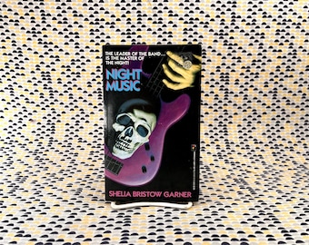 Puede incluir: Un libro de bolsillo titulado Night Music de Shelia Bristow Garner. La portada negra presenta una guitarra eléctrica morada, un cráneo humano y una mano con un anillo. El texto dice: The leader of the band... is the master of the night!