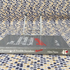 May include: A gray hardcover book titled "The Economics of Feasible Socialism" by Alec Nove. The book is on a white book stand.