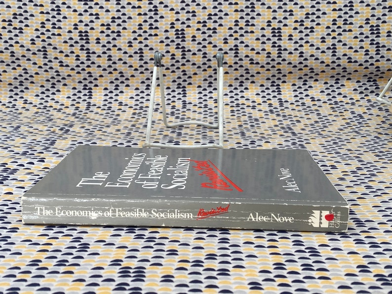 May include: A gray hardcover book titled "The Economics of Feasible Socialism" by Alec Nove. The book is on a white book stand.