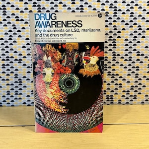 Può includere: Un libro tascabile vintage intitolato "DRUG AWARENESS" con il testo "Key documents on LSD, marijuana, and the drug culture". La copertina presenta un collage colorato con una bambola, un coniglio e altre figure. Lo sfondo è un motivo di semicerchi neri, gialli e grigi.