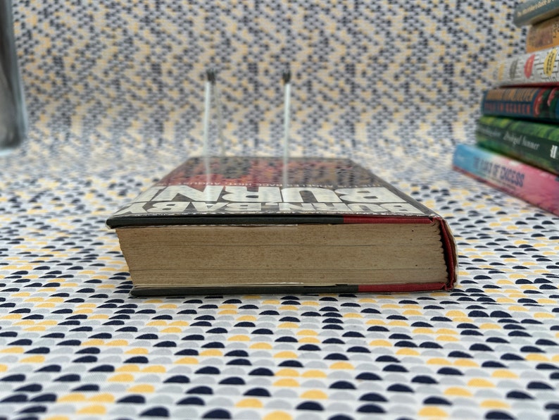 May include: A close-up of the spine of a hardcover book titled "BURN BABY BURN" by John Edgar Wideman. The book is on a patterned surface.