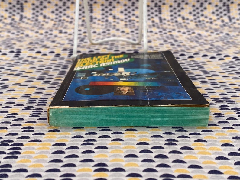 May include: A paperback book titled "The Left Hand of the Galaxy" by Isaac Asimov. The cover features a blue and black background with a colorful spectrum and a small image of a man in a white shirt.