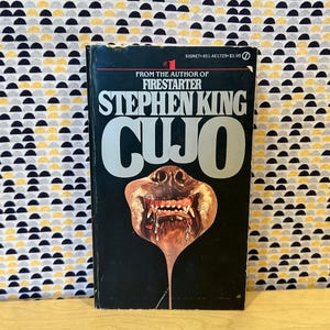 May include: A paperback book titled "CUJO" by Stephen King. The cover is black with the title in large, silver letters. The image features a snarling dog's face with exposed teeth. The book is from the author of "Firestarter".