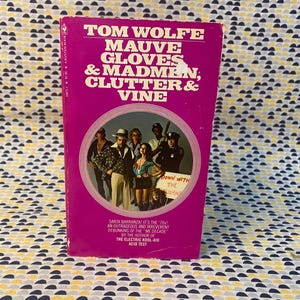 Könnte beinhalten: Ein rosa Taschenbuch mit dem Titel "Mauve Gloves & Madmen, Clutter & Vine" von Tom Wolfe. Das Cover zeigt eine Gruppe von Menschen in Kleidung aus den 1970er Jahren, mit einem Schild, auf dem "Down with the Illusion" steht.