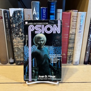 May include: A paperback book titled "Psion" by Joan D. Vinge, featuring a science fiction cover with a figure in a blue suit and a white-haired head. The book is displayed on a shelf with other books.