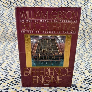 Puede incluir: Un libro de tapa dura titulado "The Difference Engine: A Novel" de William Gibson y Bruce Sterling. La portada burdeos presenta letras doradas y una imagen de una estructura industrial. Es una novela de ciencia ficción.