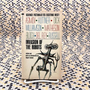 Op de afbeelding: Vintage paperback boek getiteld "Invasion of the Robots" met een retro sciencefiction illustratie van een robot. De cover toont de namen van auteurs als Asimov en Dick. Het boek staat op een witte standaard tegen een patroonachtergrond.