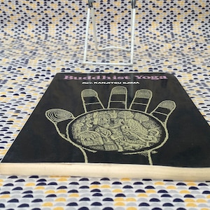 May include: A black book with the title "Buddhist Yoga" and the author "Rev. Kanjitsu Iijima". The cover features a detailed illustration of a hand with a circular design inside.