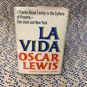 Op de afbeelding: Een boekomslag met de titel "La Vida" in blauwe en groene letters. De naam van de auteur, "Oscar Lewis", staat in rode en oranje letters onder de titel. De ondertitel van het boek is "A Puerto Rican Family in the Culture of Poverty - San Juan and New York".