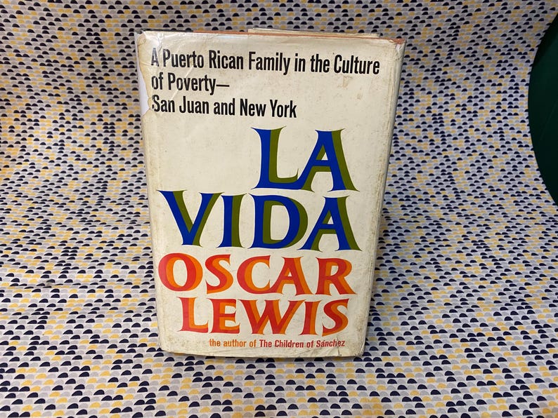 Op de afbeelding: Een boekomslag met de titel "La Vida" in blauwe en groene letters. De naam van de auteur, "Oscar Lewis", staat in rode en oranje letters onder de titel. De ondertitel van het boek is "A Puerto Rican Family in the Culture of Poverty - San Juan and New York".