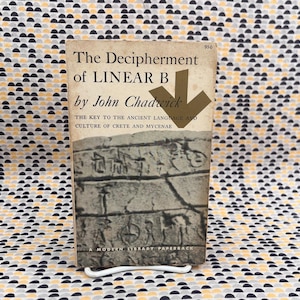 May include: A vintage paperback book titled "The Decipherment of Linear B" by John Chadwick. The cover features a stone carving and a gold arrow. The book is propped up on a white metal stand against a patterned background.