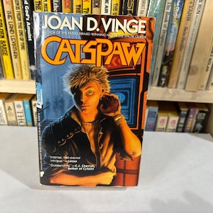 May include: A paperback book titled "CATSPAW" by Joan D. Vinge. The cover features a portrait of a person with spiky blonde hair, wearing a black jacket, and a gold watch. The background is a futuristic scene with blue and orange hues.