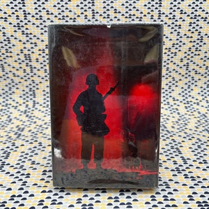 May include: A black and white image of a soldier in silhouette, holding a rifle, standing in front of a red and orange background.