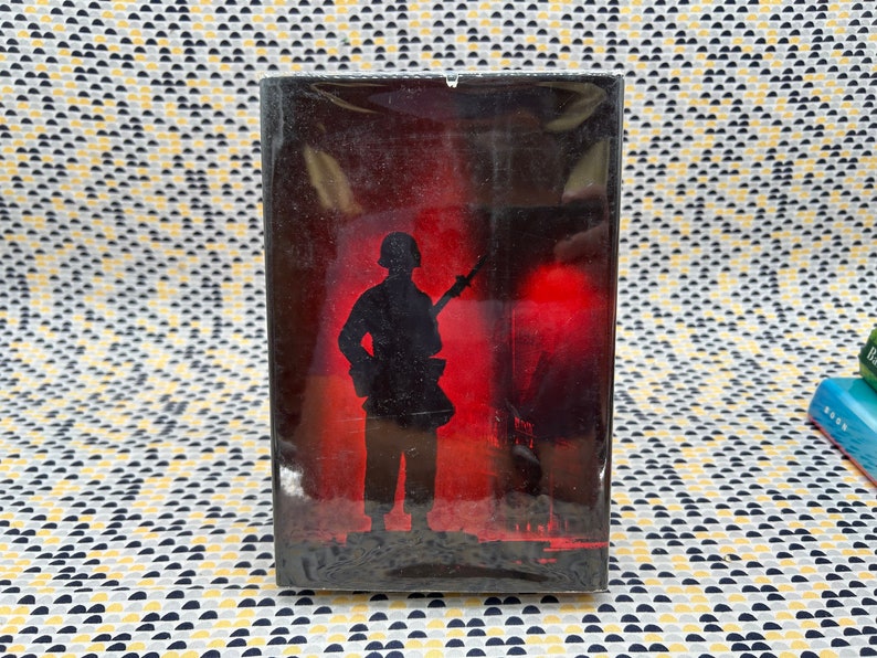 May include: A black and white image of a soldier in silhouette, holding a rifle, standing in front of a red and orange background.