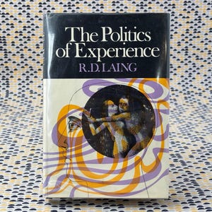 May include: A hardcover book titled "The Politics of Experience" by R.D. Laing. The cover features a black top section with white text and a lower section with a surreal illustration in orange, purple, and beige.