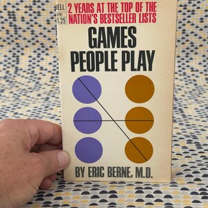 Może przedstawiać: Książka w miękkiej oprawie w stylu vintage zatytułowana "Games People Play" autorstwa Erica Berne, M.D. Okładka zawiera tytuł pogrubionymi czarnymi literami, z grafiką połączonych kół w kolorze fioletowym i brązowym. Książka to wydanie Dell.