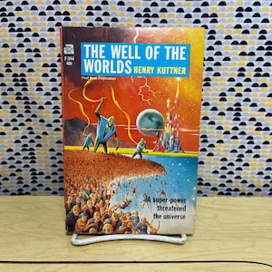 Op de afbeelding: Vintage paperback boekcover voor "The Well of the Worlds" van Henry Kuttner. De coverkunst toont buitenaardse figuren, een futuristische stad en een menigte mensen die in een blauwe afgrond vallen. De titel staat in een blauwe doos.