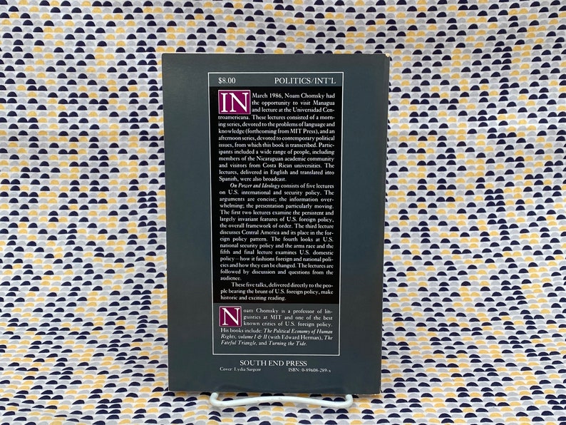 Op de afbeelding: Een zwart-witte boekomslag met de titel "Politics/Int'l" en de naam van de auteur "Noam Chomsky". De boekomslag bevat een korte beschrijving van de inhoud van het boek, inclusief een samenvatting van de lezingen en de achtergrond van de auteur.