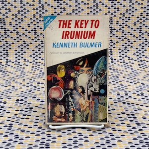 Może przedstawiać: Antyczna książka w miękkiej oprawie zatytułowana "The Key to Irinium" autorstwa Kennetha Bulmera. Okładka przedstawia kolorowy kolaż obrazów o tematyce kosmicznej, w tym planety, gwiazdy i futurystyczne miasto.