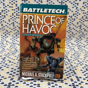 May include: A paperback book titled "Battletech Prince of Havoc" with a cover featuring a dark green robot and a blue robot. The book is by Michael A. Stackpole and is part of the "Twilight of the Clans VII" series.