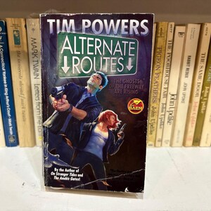 May include: A paperback book titled "Alternate Routes" by Tim Powers. The cover features two figures holding guns, with the text "The Ghosts of the Freeway Are Rising." The book is displayed on a shelf with other books.
