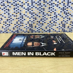 Puede incluir: Un libro de bolsillo negro con una foto de tres hombres con trajes negros en la portada. El t&iacute;tulo "Men in Black" est&aacute; impreso en blanco en el lomo. El libro forma parte de la serie "Movie Tie-In".