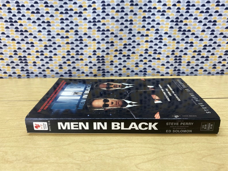 Puede incluir: Un libro de bolsillo negro con una foto de tres hombres con trajes negros en la portada. El t&iacute;tulo "Men in Black" est&aacute; impreso en blanco en el lomo. El libro forma parte de la serie "Movie Tie-In".