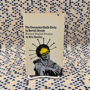 May include: A vintage paperback book titled "The Caucasian Chalk Circle" by Bertolt Brecht, Revised English Version by Eric Bentley. The cover features a stylized illustration of a figure with a yellow mask and radiating lines.