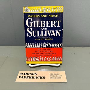 May include: A paperback book titled Words and Music Gilbert and Sullivan with an introduction by Alan Pitt Robbins. The cover features yellow, blue, and red sections with black and white illustrations of figures and the text The Mikado, H.M.S. Pinafore, Pirates of Penzance.