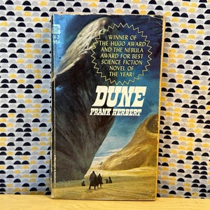 May include: Vintage paperback book cover for "Dune" by Frank Herbert. The cover features a large, dark, worm-like creature and a desert landscape. Text includes the title "Dune", the author's name, and awards won, including the Hugo Award.