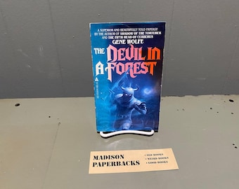 Può includere: Un libro tascabile intitolato The Devil in a Forest di Gene Wolfe con una copertina blu che presenta l'illustrazione di un guerriero cornuto. Sotto il libro c'è una carta rettangolare con il testo Madison Paperbacks, Old Books, Weird Books, Good Books.