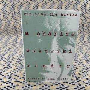 Puede incluir: Un libro de bolsillo titulado "Run With The Hunted: A Charles Bukowski Reader". La portada es verde claro con texto rojo y una imagen en escala de grises del rostro de un hombre. Editado por John Martin.